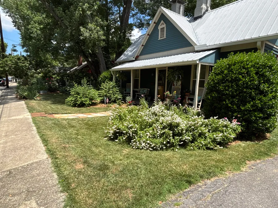 Street-side view of a residential front yard with lawn, flowering plants, shrubs, and a front porch.